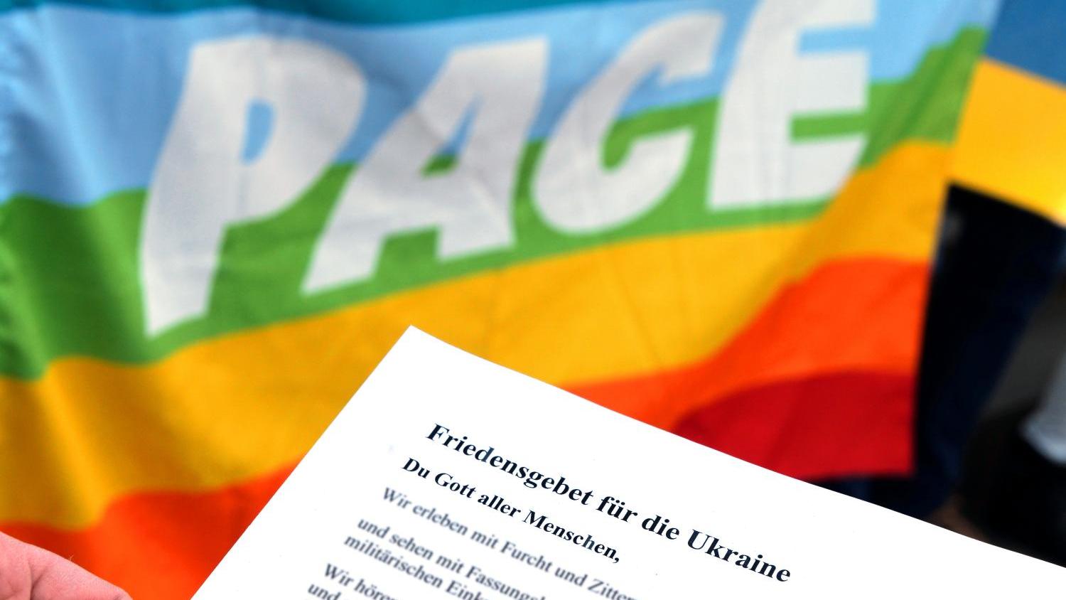Das Bild zeigt die regenbogenfarbene Friedensflagge mit dem Wort 'pace' und einem davorgehaltenen Blatt Papier mit einem Friedensgebet für die Ukraine