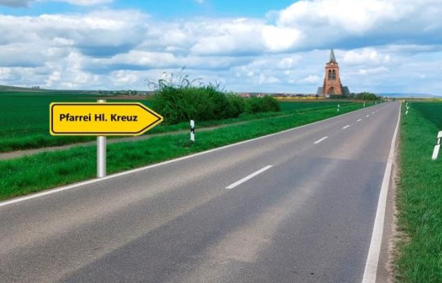 Das Bild zeigt eine Wiesenlandschaft mit einem Kirchturm im Hintergrund und einem Straßenschild mit der Aufschrift Heilig Kreuz