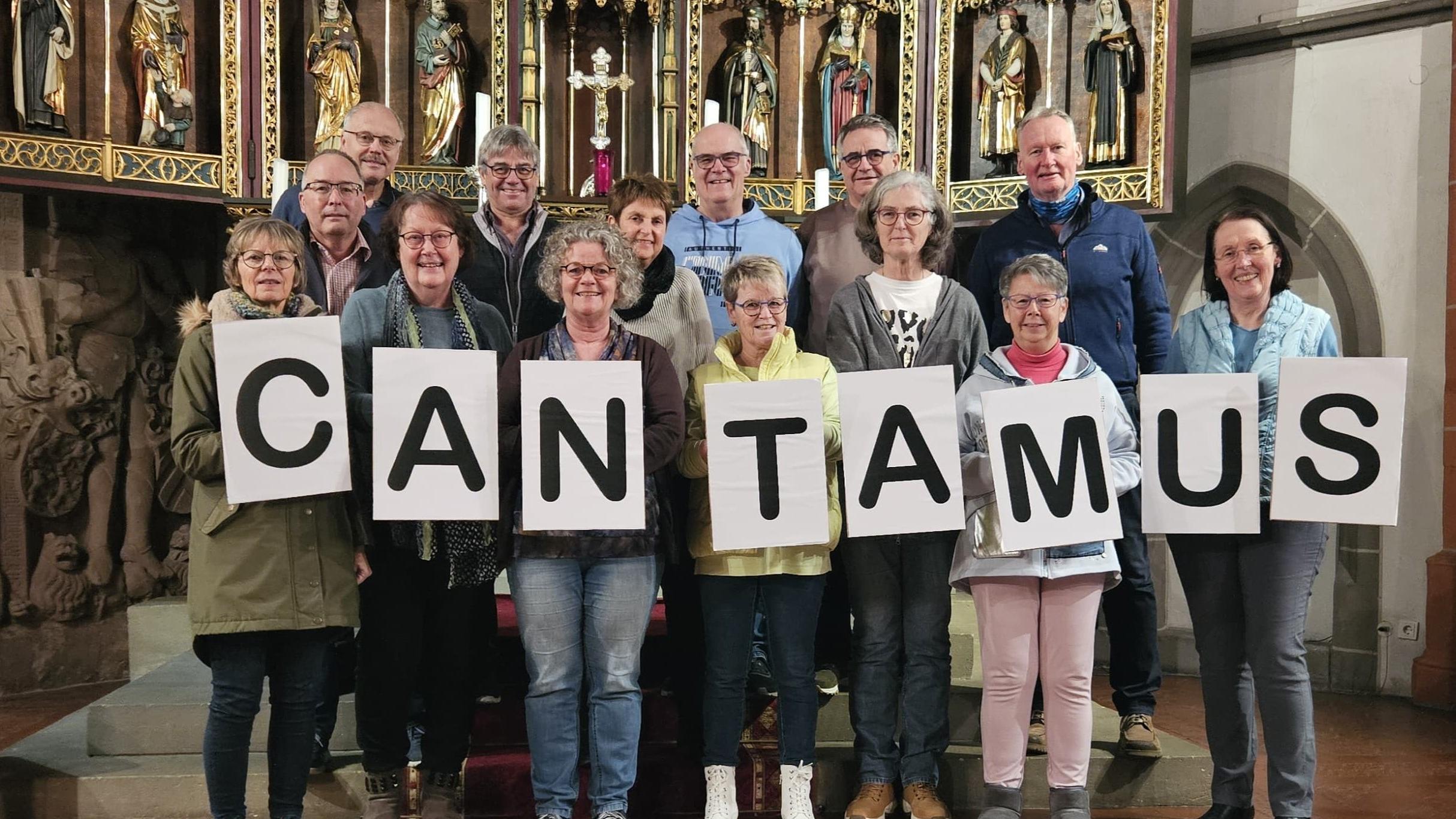 Bild mit den Chormitgliedern des Chores Cantamus im Chorraum der St.-Nikolaus-Kirche. Die Sänger halten große Buchstaben in der Hand, die das Wort 'Cantamus' bilden.