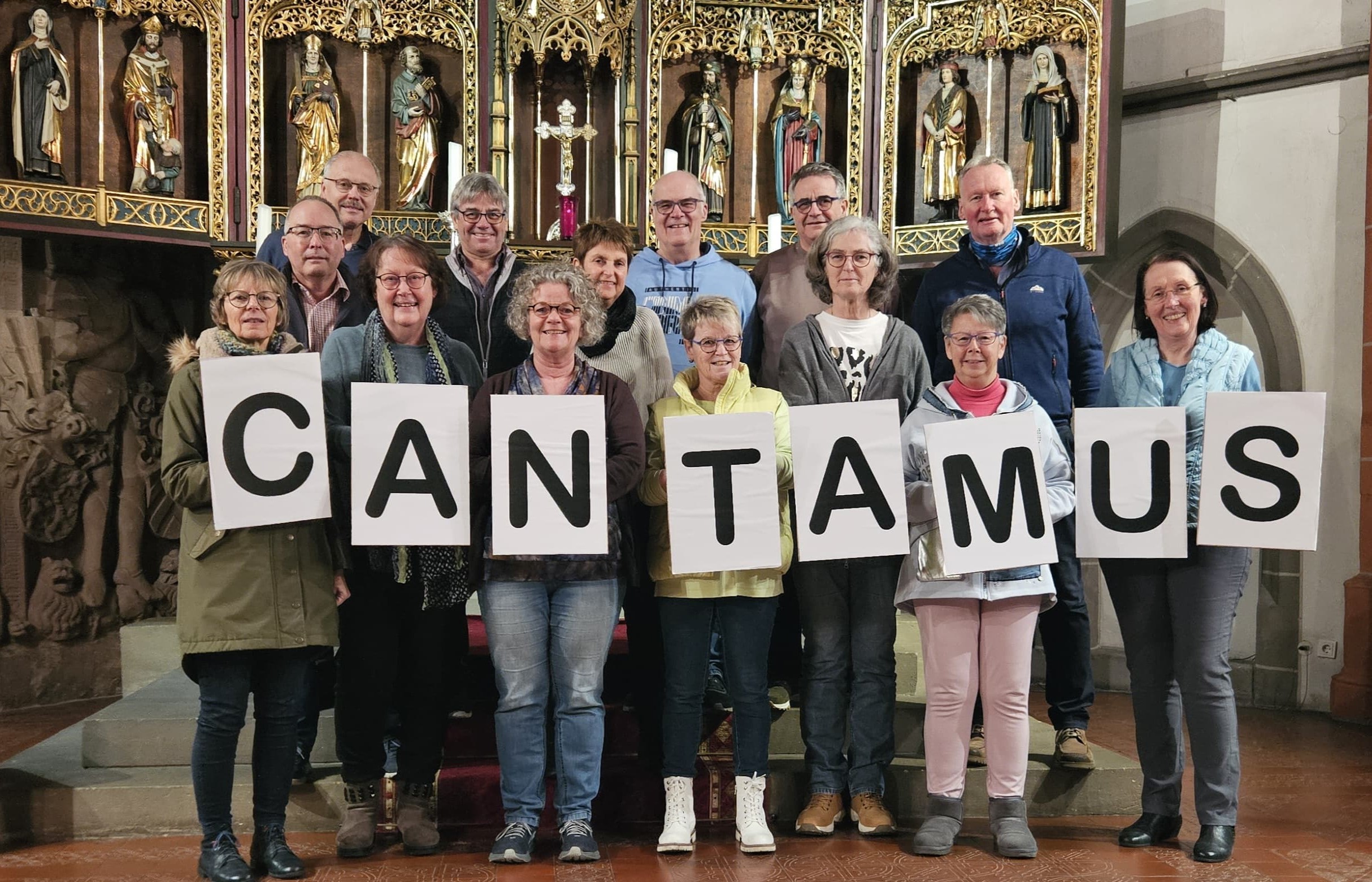 Bild mit den Chormitgliedern des Chores Cantamus im Chorraum der St.-Nikolaus-Kirche. Die Sänger halten große Buchstaben in der Hand, die das Wort 'Cantamus' bilden.