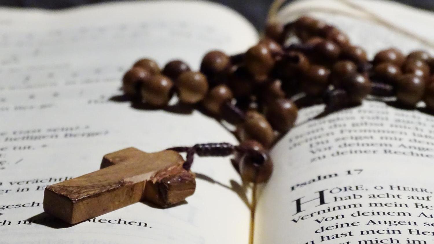 Holzrosenkranz mit Kreuz liegt auf aufgeschlagenem Buch.